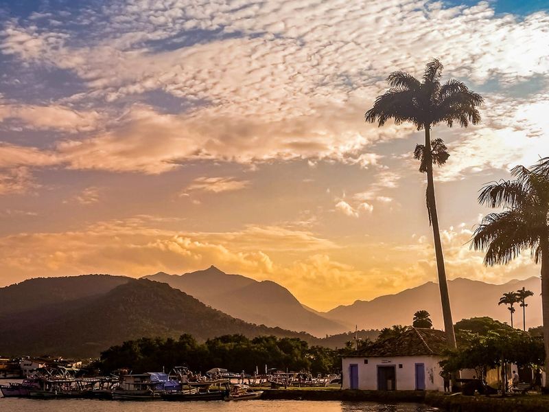 Sunset over a coastal town with mountains in the background, palm trees, boats docked by the shore, and a vibrant sky with scattered clouds.