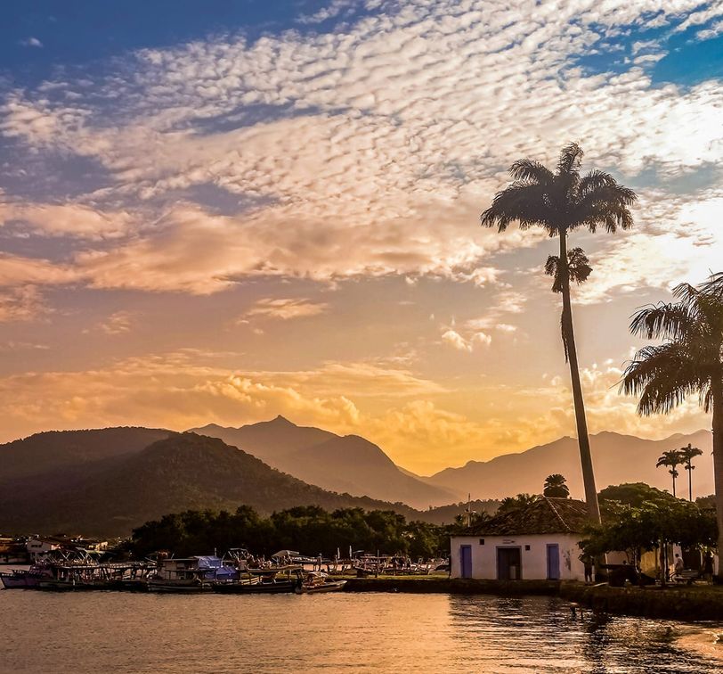 Sunset over a coastal town with mountains in the background, palm trees, boats docked by the shore, and a vibrant sky with scattered clouds.