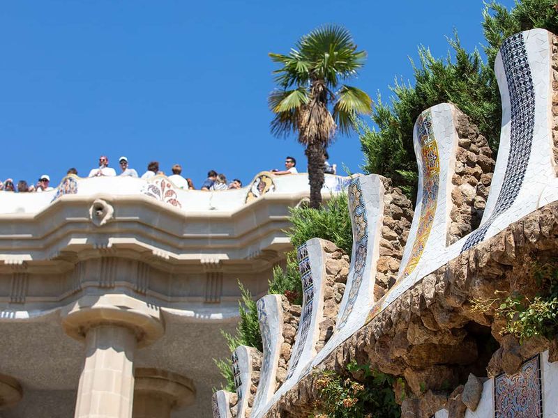 A view of Park Güell's vibrant, mosaic-adorned terrace, with visitors observing and a palm tree against a clear blue sky in Barcelona.