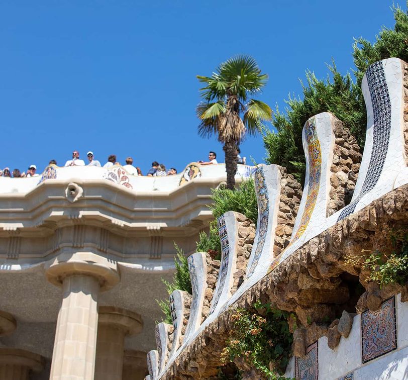 A view of Park Güell's vibrant, mosaic-adorned terrace, with visitors observing and a palm tree against a clear blue sky in Barcelona.