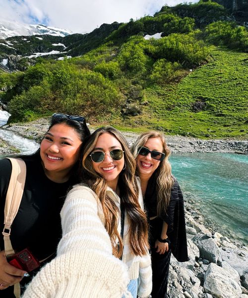 Three women taking a selfie by a turquoise river, with a lush green hillside and snow-capped mountains in the background.