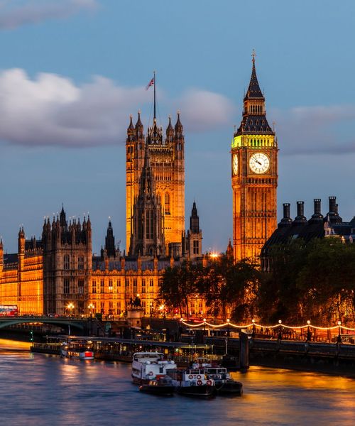 Night view of the illuminated Palace of Westminster and Big Ben beside the River Thames, with boats in the foreground and a cloudy sky above.