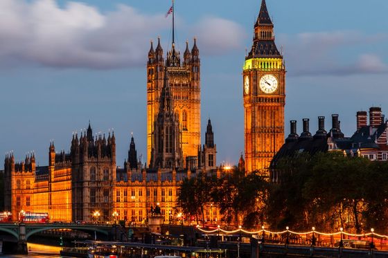 Night view of the illuminated Palace of Westminster and Big Ben beside the River Thames, with boats in the foreground and a cloudy sky above.