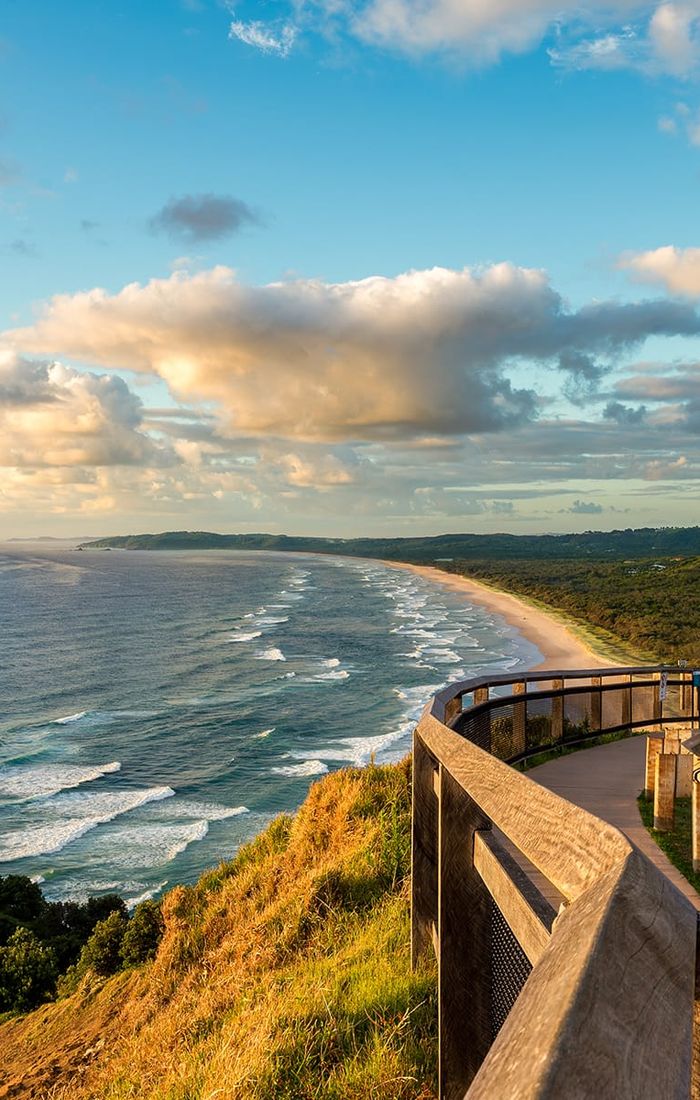 Scenic coastal view from a wooden lookout, overlooking a sandy beach, lush greenery, and a vast ocean under a partly cloudy sky.