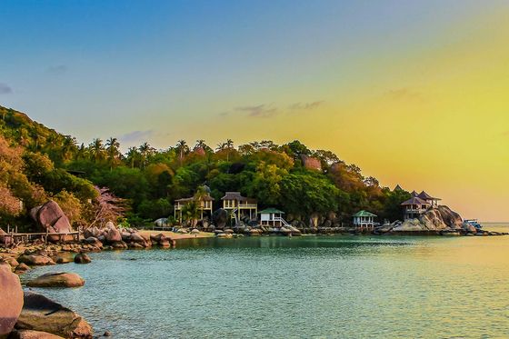 Scenic view of a tropical coastline at sunset, featuring rocky shores, lush greenery, and a few buildings nestled among the trees.