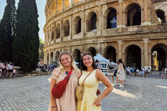 Two women in summer dresses stand smiling in front of the Colosseum in Rome, with tourists and a blue sky in the background.