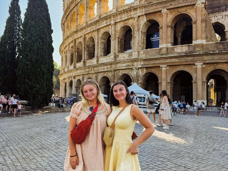 Two women in summer dresses stand smiling in front of the Colosseum in Rome, with tourists and a blue sky in the background.
