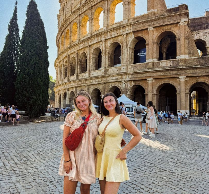 Two women in summer dresses stand smiling in front of the Colosseum in Rome, with tourists and a blue sky in the background.