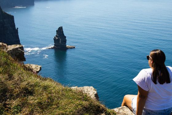 A woman in sunglasses sits on a grassy cliff edge overlooking a vast ocean with distant cliffs and a lone sea stack on a clear day.