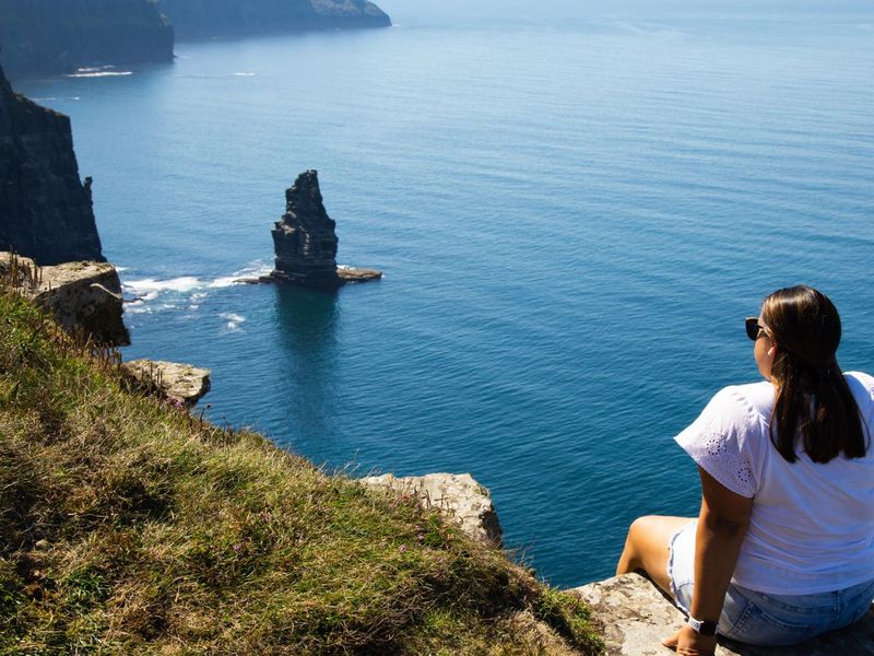 A woman in sunglasses sits on a grassy cliff edge overlooking a vast ocean with distant cliffs and a lone sea stack on a clear day.