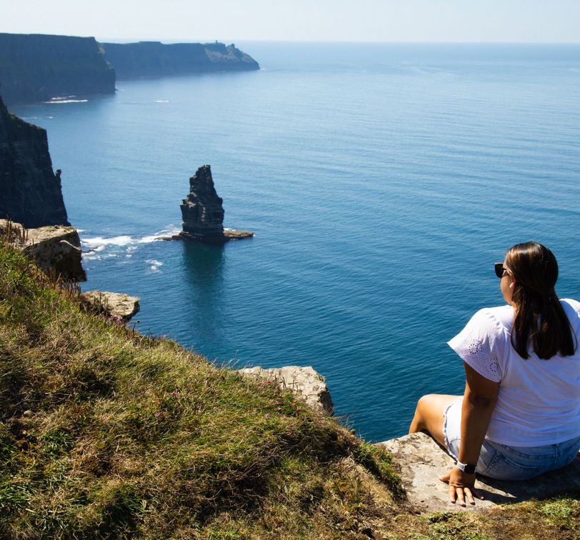 A woman in sunglasses sits on a grassy cliff edge overlooking a vast ocean with distant cliffs and a lone sea stack on a clear day.