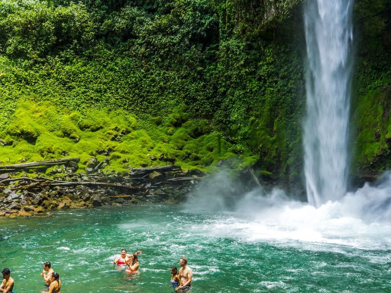 People swimming in a clear, turquoise pool at the base of a lush, green waterfall surrounded by dense foliage.