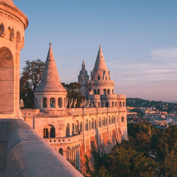 A castle-like structure with conical towers bathed in warm sunlight, surrounded by trees, overlooking a distant cityscape under a clear sky.
