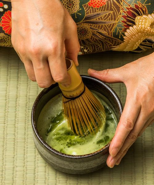 Hands whisking matcha green tea in a bowl on a tatami mat, with a floral kimono pattern visible in the background.