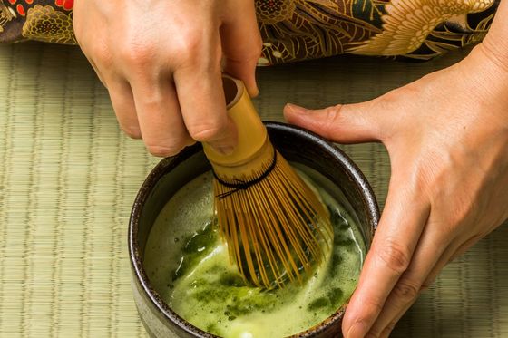 Hands whisking matcha green tea in a bowl on a tatami mat, with a floral kimono pattern visible in the background.