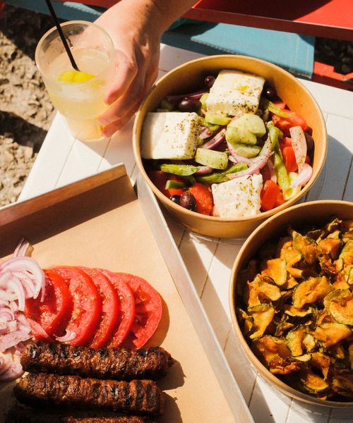 A hand holds a lemonade next to dishes of Greek salad with feta, fried zucchini chips, and a platter of grilled sausages, tomatoes, and onions.