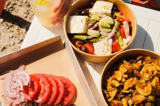 A hand holds a lemonade next to dishes of Greek salad with feta, fried zucchini chips, and a platter of grilled sausages, tomatoes, and onions.