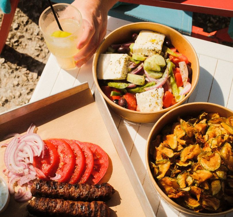 A hand holds a lemonade next to dishes of Greek salad with feta, fried zucchini chips, and a platter of grilled sausages, tomatoes, and onions.