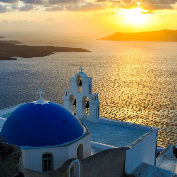 A blue-domed church with bell towers overlooks a golden sunset on the sea, set against a silhouette of islands.
