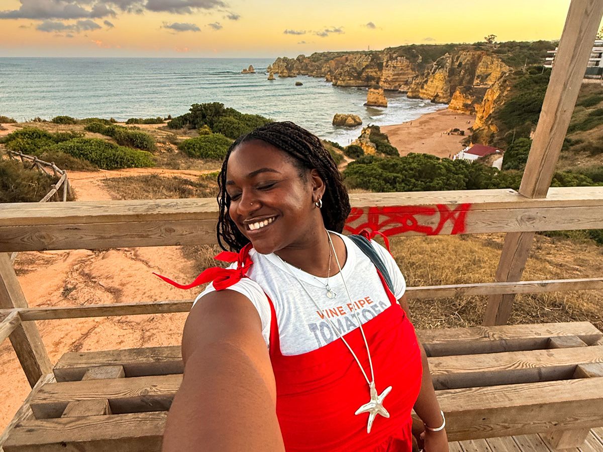Woman in red dress takes a selfie on a wooden deck overlooking a coastal landscape with cliffs and ocean at sunset.