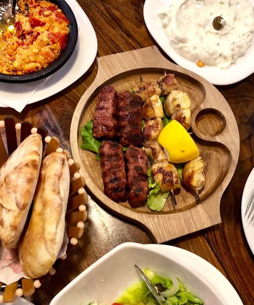 A wooden platter with grilled meats and lemon, surrounded by bread, salad, yogurt dip, and a spicy dish on a wooden table.