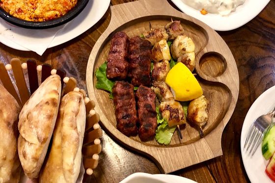 A wooden platter with grilled meats and lemon, surrounded by bread, salad, yogurt dip, and a spicy dish on a wooden table.