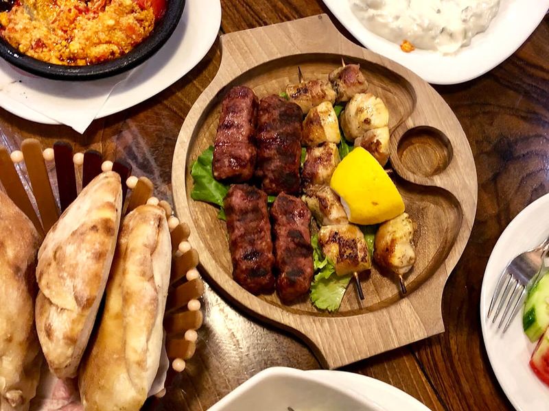 A wooden platter with grilled meats and lemon, surrounded by bread, salad, yogurt dip, and a spicy dish on a wooden table.