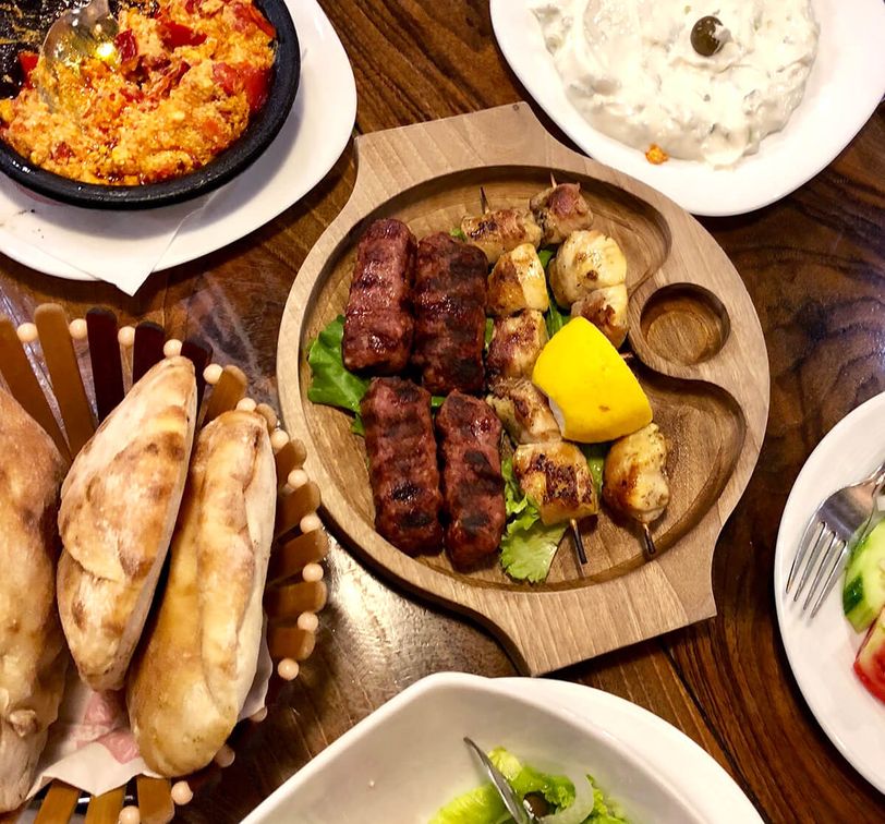 A wooden platter with grilled meats and lemon, surrounded by bread, salad, yogurt dip, and a spicy dish on a wooden table.
