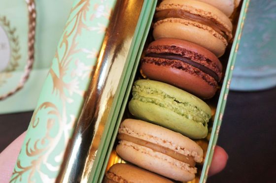 A hand holds an open, decorative box containing six colorful macarons in shades of beige, brown, and green, with a gold interior.