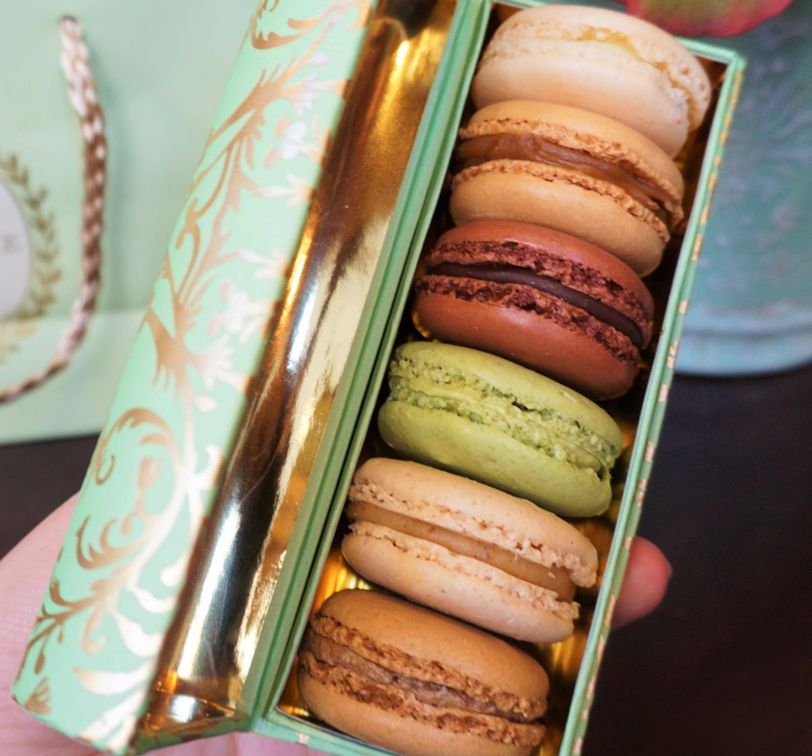 A hand holds an open, decorative box containing six colorful macarons in shades of beige, brown, and green, with a gold interior.