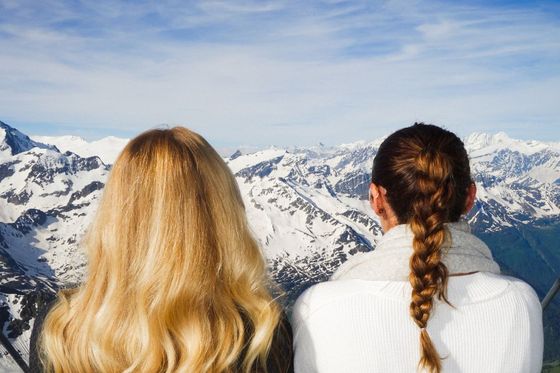 Two women with long hair admire a breathtaking view of snow-capped mountains under a vibrant blue sky.