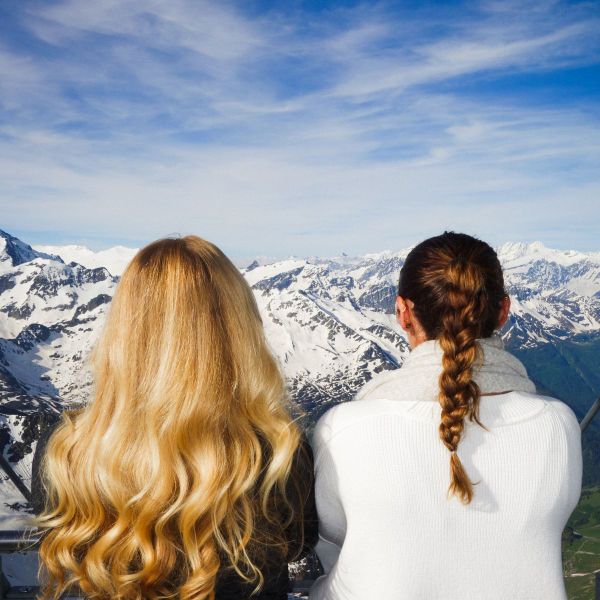 Two women with long hair admire a breathtaking view of snow-capped mountains under a vibrant blue sky.