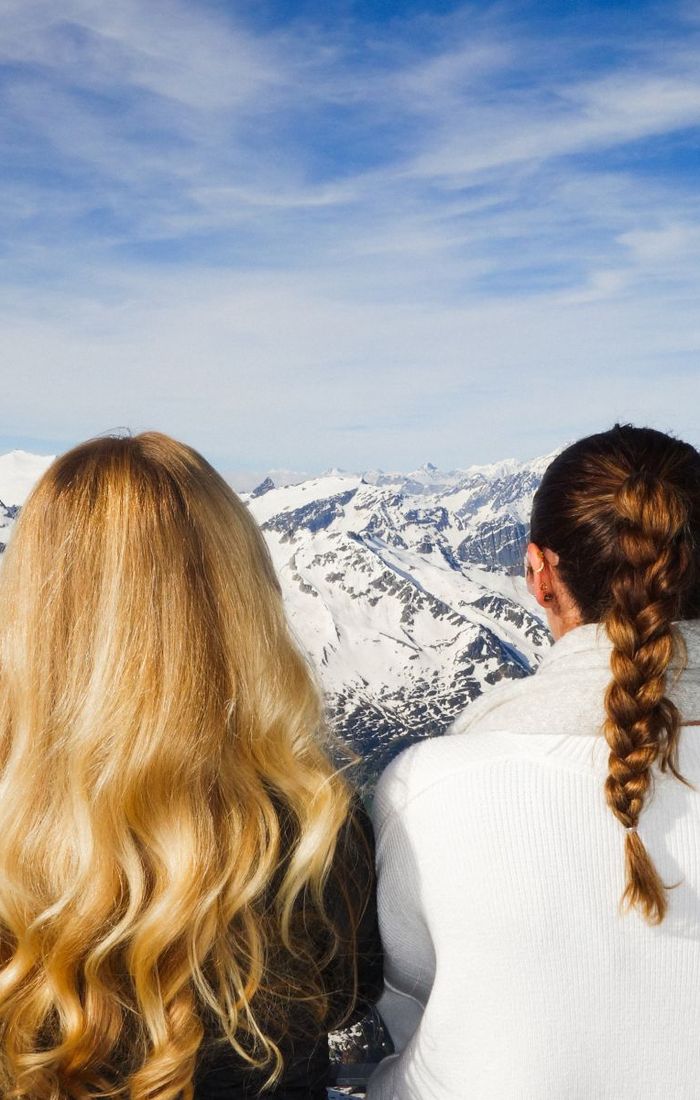 Two women with long hair admire a breathtaking view of snow-capped mountains under a vibrant blue sky.