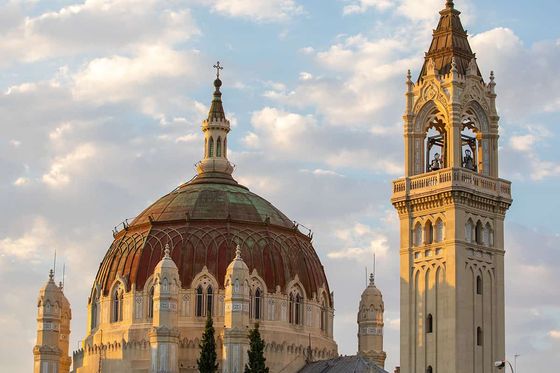 A grand church with a large dome and ornate bell tower under a partly cloudy sky, surrounded by trees.
