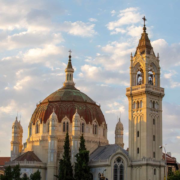A grand church with a large dome and ornate bell tower under a partly cloudy sky, surrounded by trees.