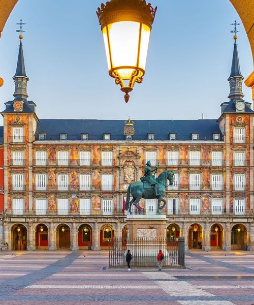 A historical plaza with a central equestrian statue and ornate building, framed by an archway with a street lamp above.