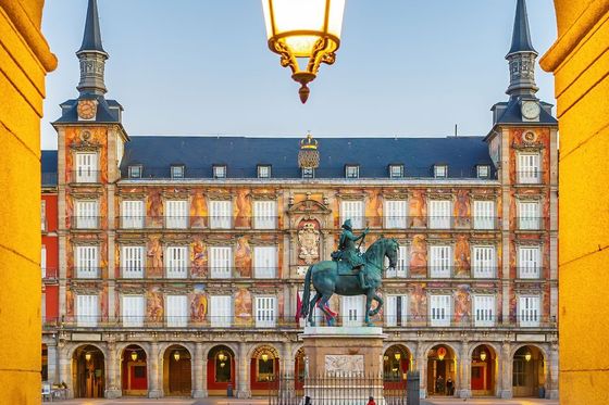 A historical plaza with a central equestrian statue and ornate building, framed by an archway with a street lamp above.