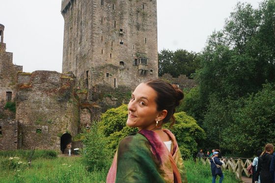 A person smiling and wrapped in a colorful shawl stands in front of an ancient stone castle surrounded by lush greenery.
