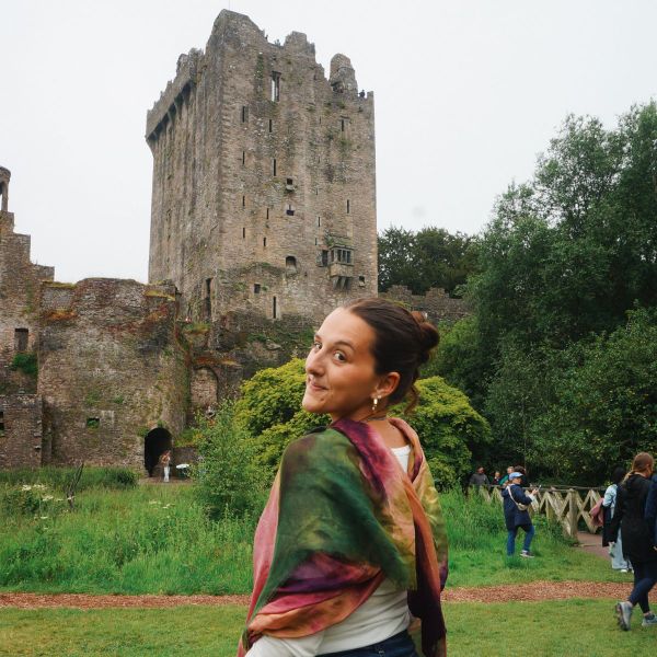 A person smiling and wrapped in a colorful shawl stands in front of an ancient stone castle surrounded by lush greenery.