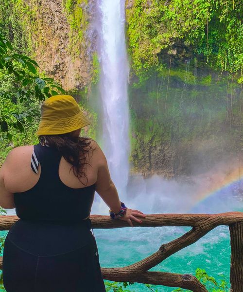 A person in a yellow hat and black outfit stands on a wooden lookout, gazing at a waterfall and rainbow in a lush, green environment.