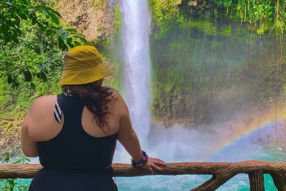 A person in a yellow hat and black outfit stands on a wooden lookout, gazing at a waterfall and rainbow in a lush, green environment.
