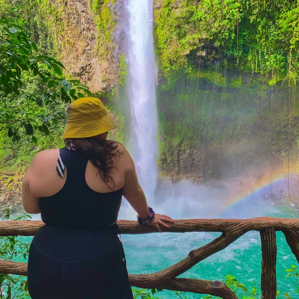 A person in a yellow hat and black outfit stands on a wooden lookout, gazing at a waterfall and rainbow in a lush, green environment.
