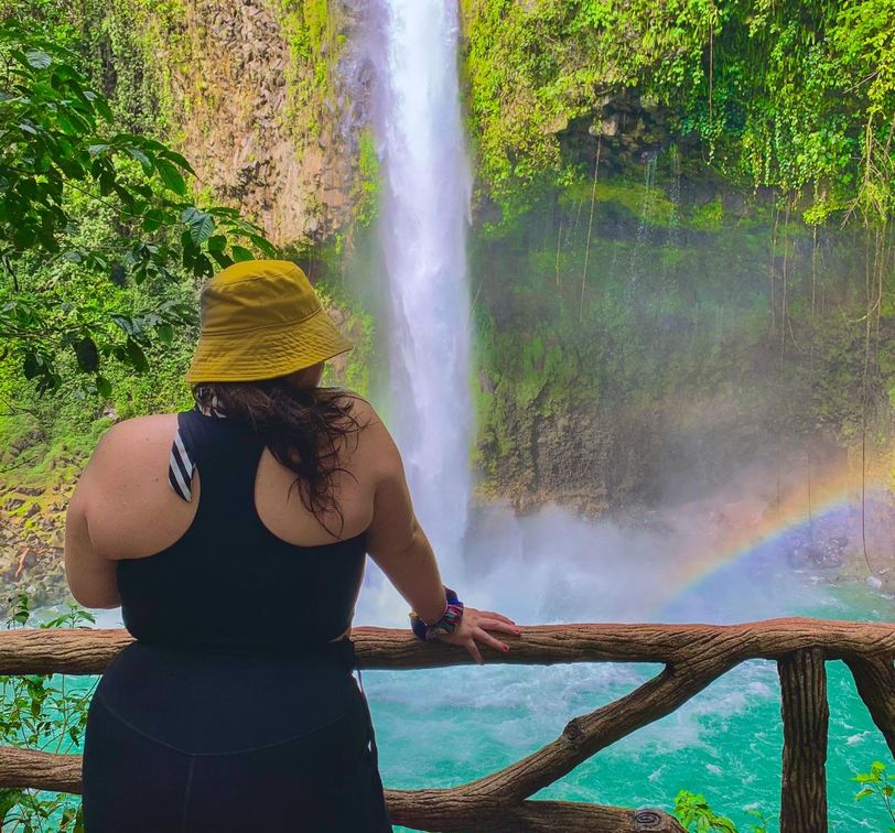 A person in a yellow hat and black outfit stands on a wooden lookout, gazing at a waterfall and rainbow in a lush, green environment.