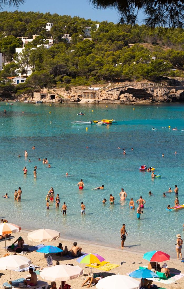 Beachgoers enjoy swimming and sunbathing on a sandy beach with turquoise water, surrounded by trees and rocky cliffs. Boats are anchored offshore.
