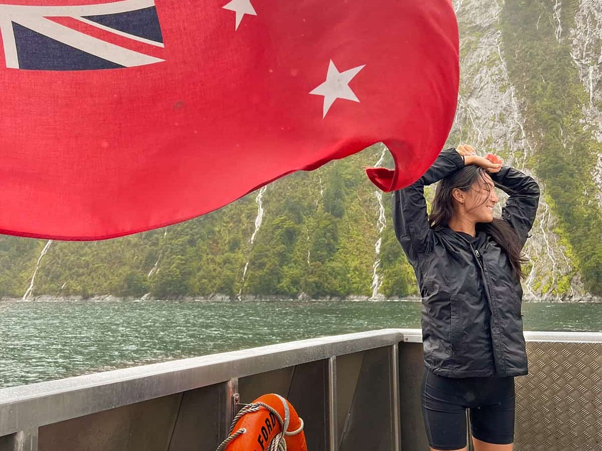A person stands on a boat in front of a red flag, with lush green cliffs and a misty sky in the background.