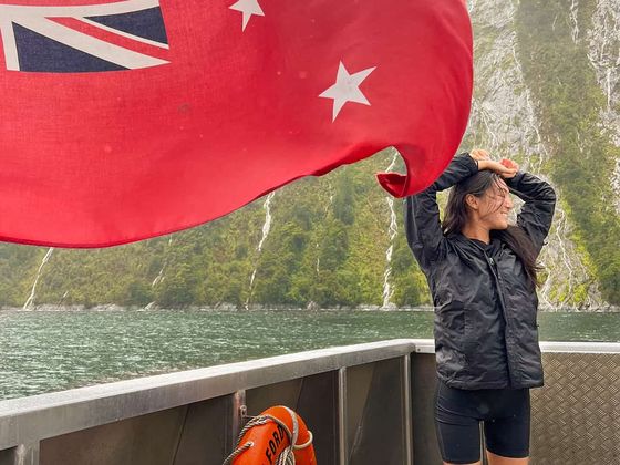 A person stands on a boat in front of a red flag, with lush green cliffs and a misty sky in the background.