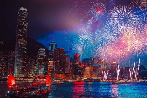 A vibrant fireworks display lights up the night sky over a city skyline with a traditional boat on the water, reflecting colorful lights.