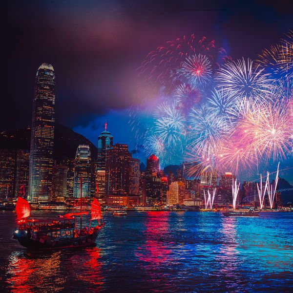 A vibrant fireworks display lights up the night sky over a city skyline with a traditional boat on the water, reflecting colorful lights.