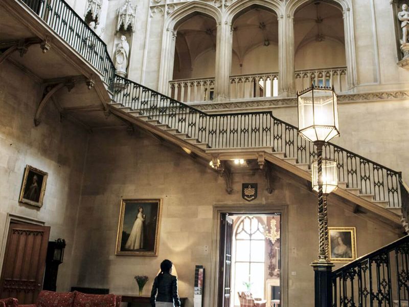 A grand staircase in a historic building with arched windows, paintings, ornate railings, and a person walking below.
