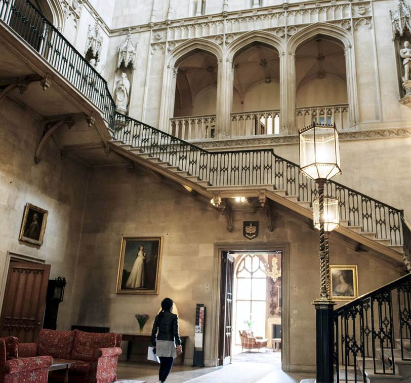 A grand staircase in a historic building with arched windows, paintings, ornate railings, and a person walking below.